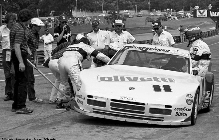 The #11 Porsche 944 in the pits with a driver climbing in and the crew working
