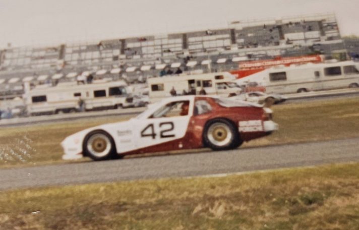 The #42 Porsche 944 in red and white on a circuit in front of a large grandstand